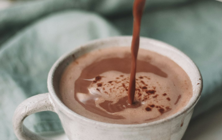 Hot cocoa being poured into a white ceramic mug with a light green fabric in the background